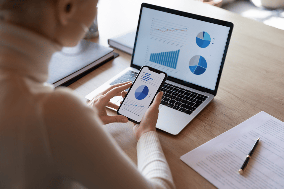 A woman looks at her phone and computer, looking at financial charts
